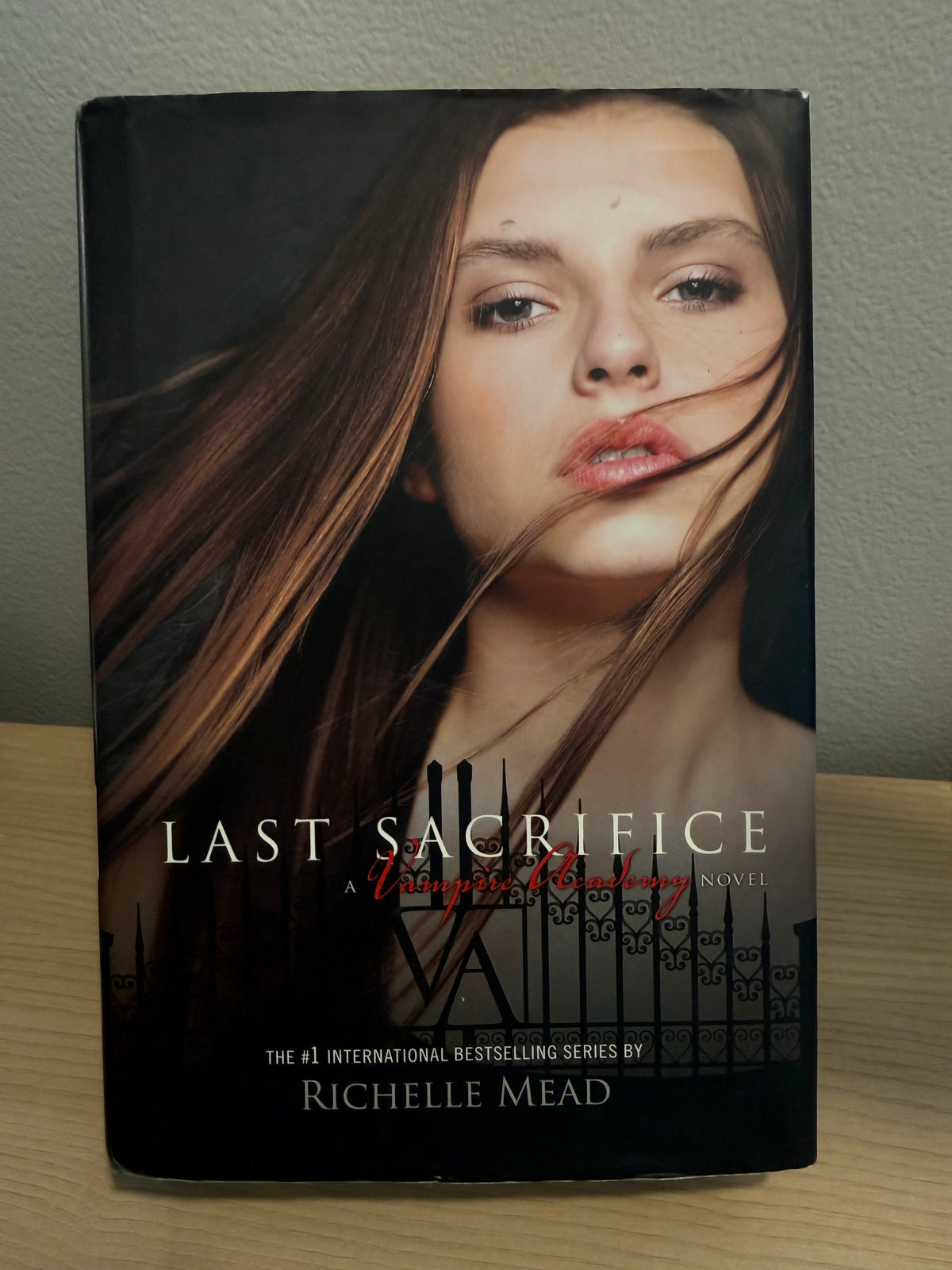 The book 'Last Sacrifice' by Richelle Mead, featuring a portrait of a woman with flowing brown hair on the cover, standing on a light wood surface against a gray wall.