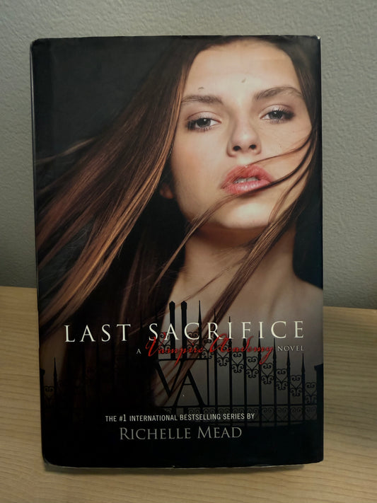 The book 'Last Sacrifice' by Richelle Mead, featuring a portrait of a woman with flowing brown hair on the cover, standing on a light wood surface against a gray wall.