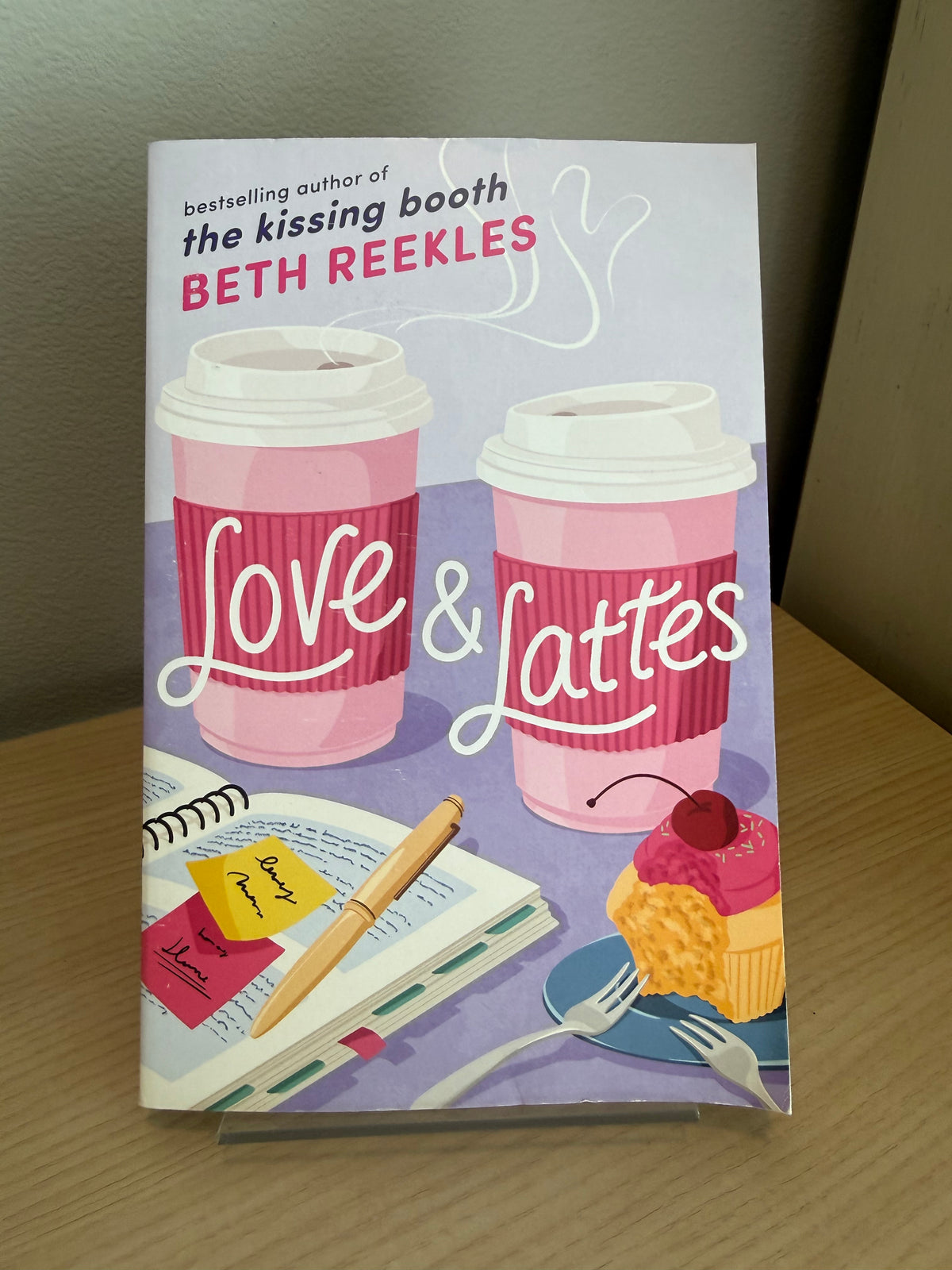 The book 'Love & Lattes' by Beth Reekles, featuring a colorful cover illustration of two pink coffee cups, a notebook, a pen, and a cupcake, displayed on a clear acrylic stand on a wooden table.
