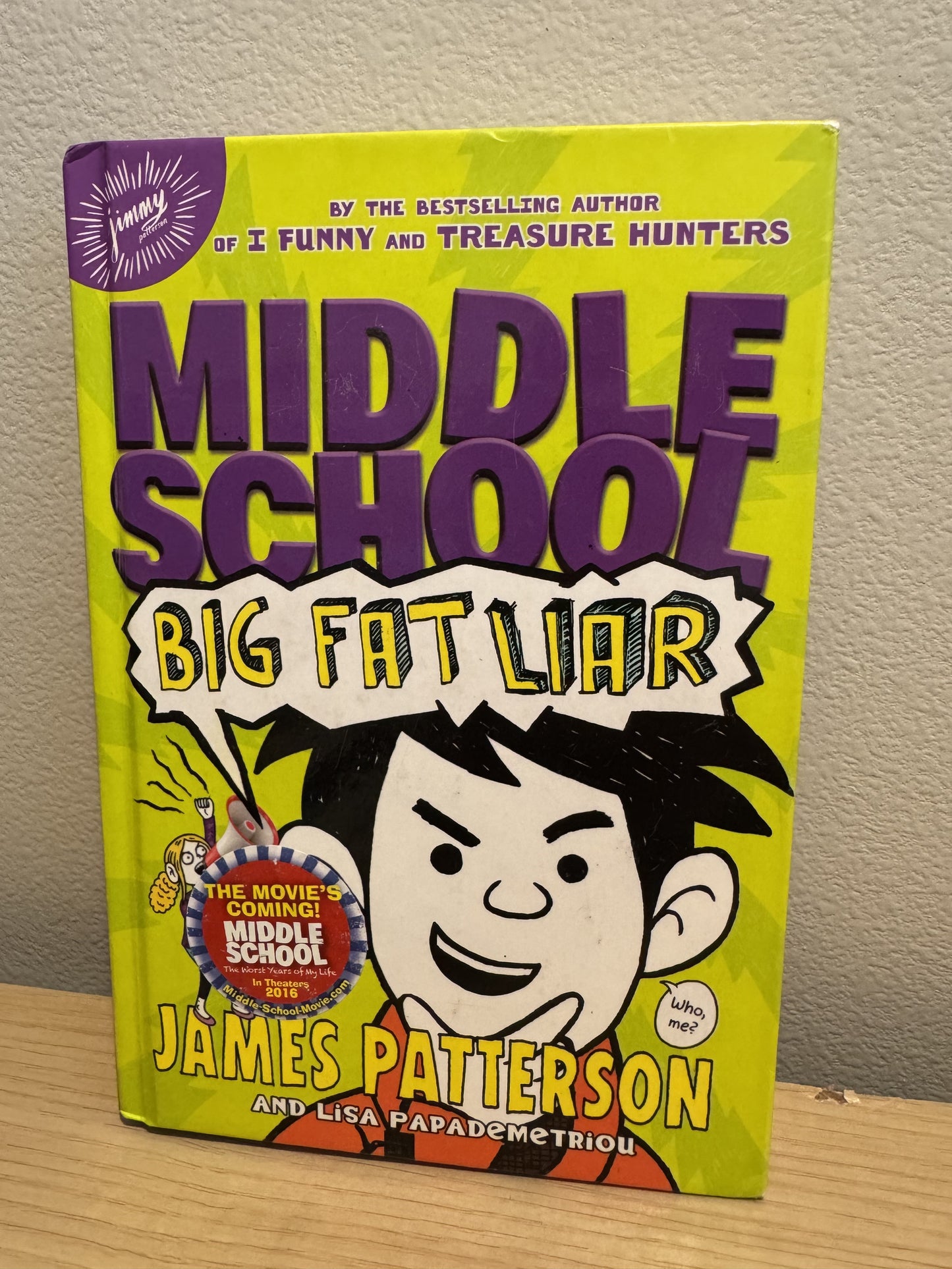 The front cover of the children's book 'Middle School: Big Fat Liar' by James Patterson and Lisa Papademetriou, featuring a bright lime green background, bold purple title text, and an illustrated cartoon boy with black hair and a white face. The book is standing upright on a light wood surface against a beige wall.