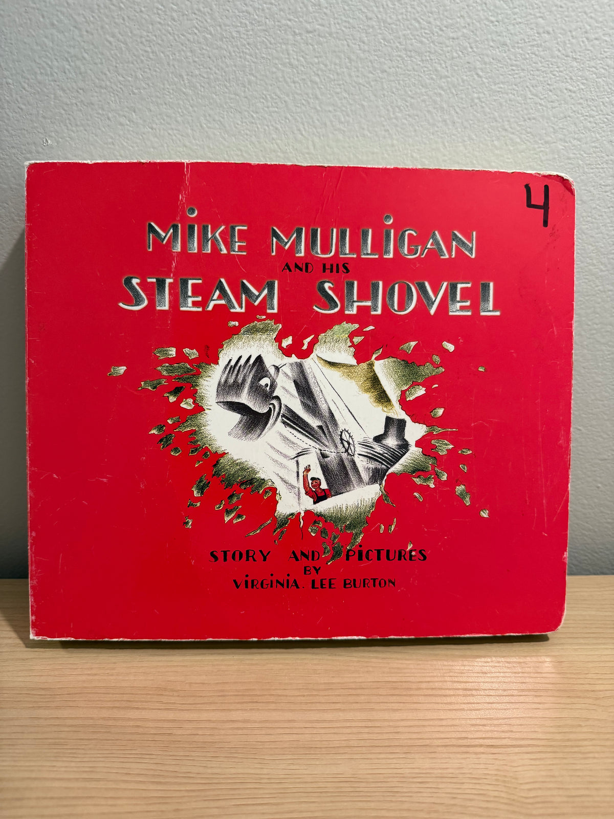 Vintage children's book titled "Mike Mulligan and His Steam Shovel" by Virginia Lee Burton, featuring a bright red cover with a central illustration of a steam shovel and the title in large white letters, photographed standing on a light wood surface against a gray wall.