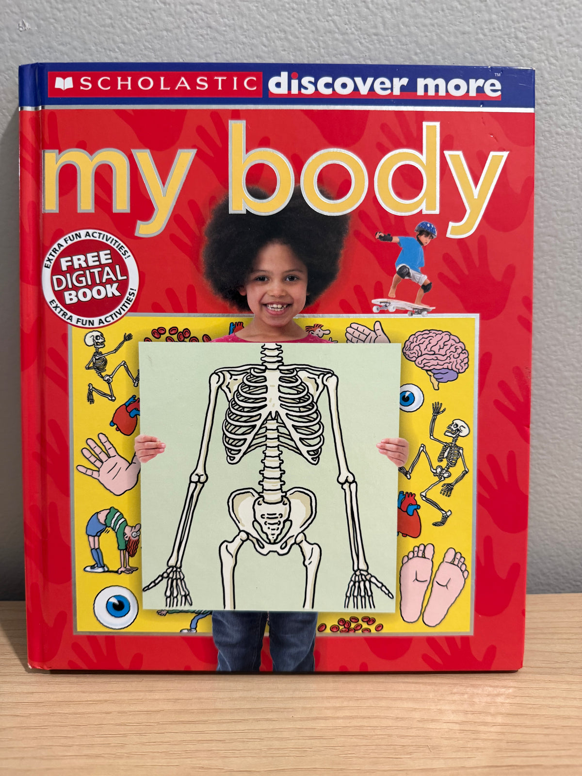 A child with an afro hairstyle stands behind a large red Scholastic 'discover more my body' book, holding a printed sheet with a black-and-white human skeleton illustration. The book cover features colorful illustrations and a 'FREE DIGITAL BOOK' badge.