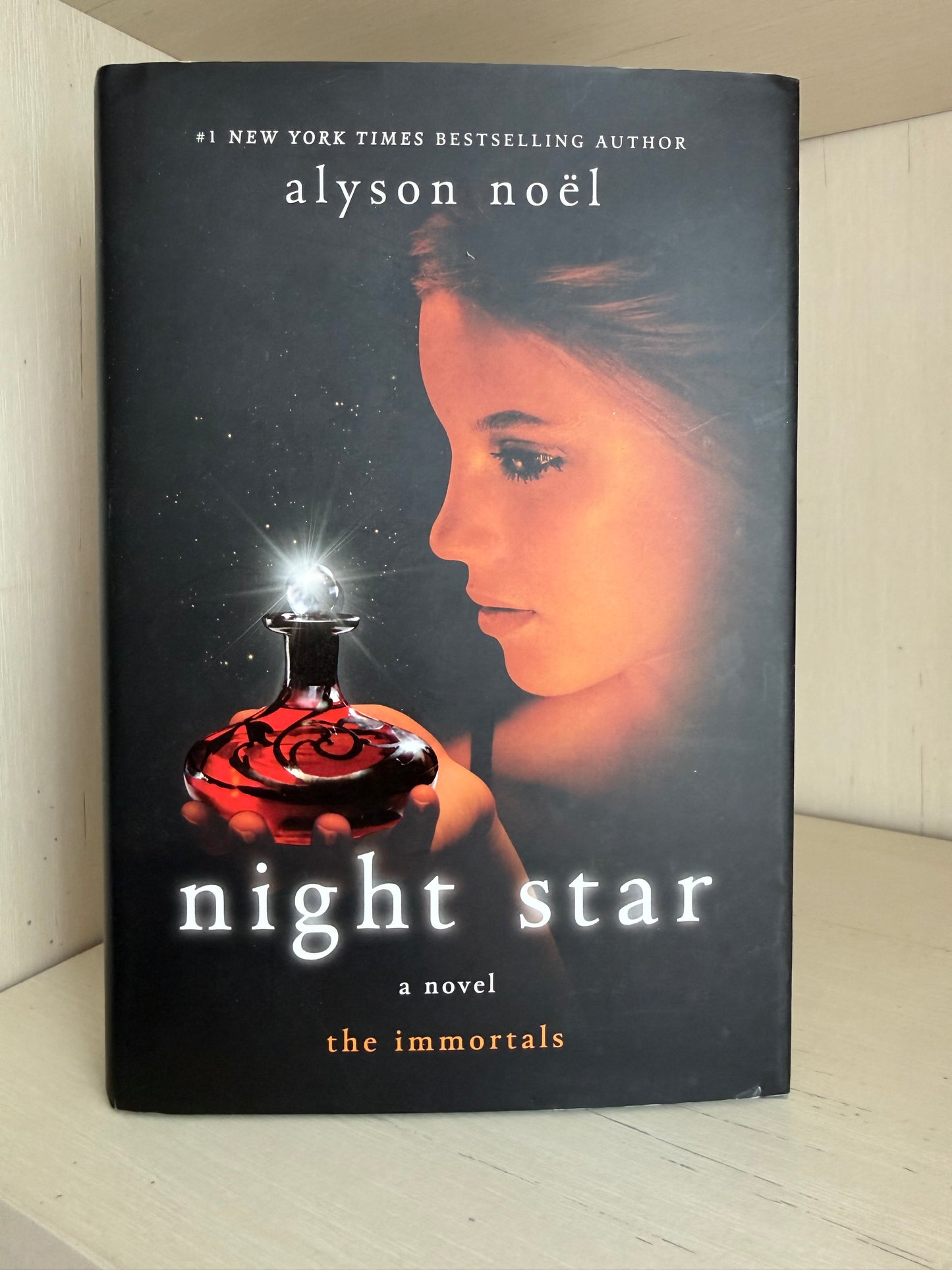 A hardcover copy of the novel 'Night Star' by Alyson Noel, leaning against a light-colored wooden surface. The book cover is predominantly black, featuring a glowing orange-toned portrait of a woman and a dark red glass bottle with a bright light source.
