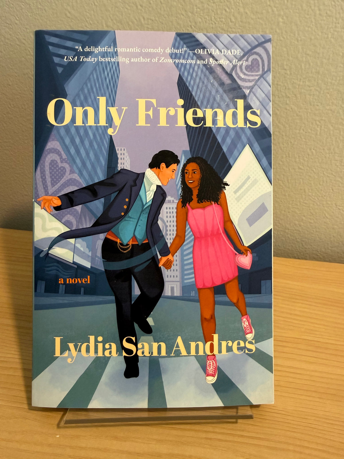 The book cover of 'Only Friends' by Lydia San Andres, displayed upright on a clear plastic stand on a wooden table against a beige wall.