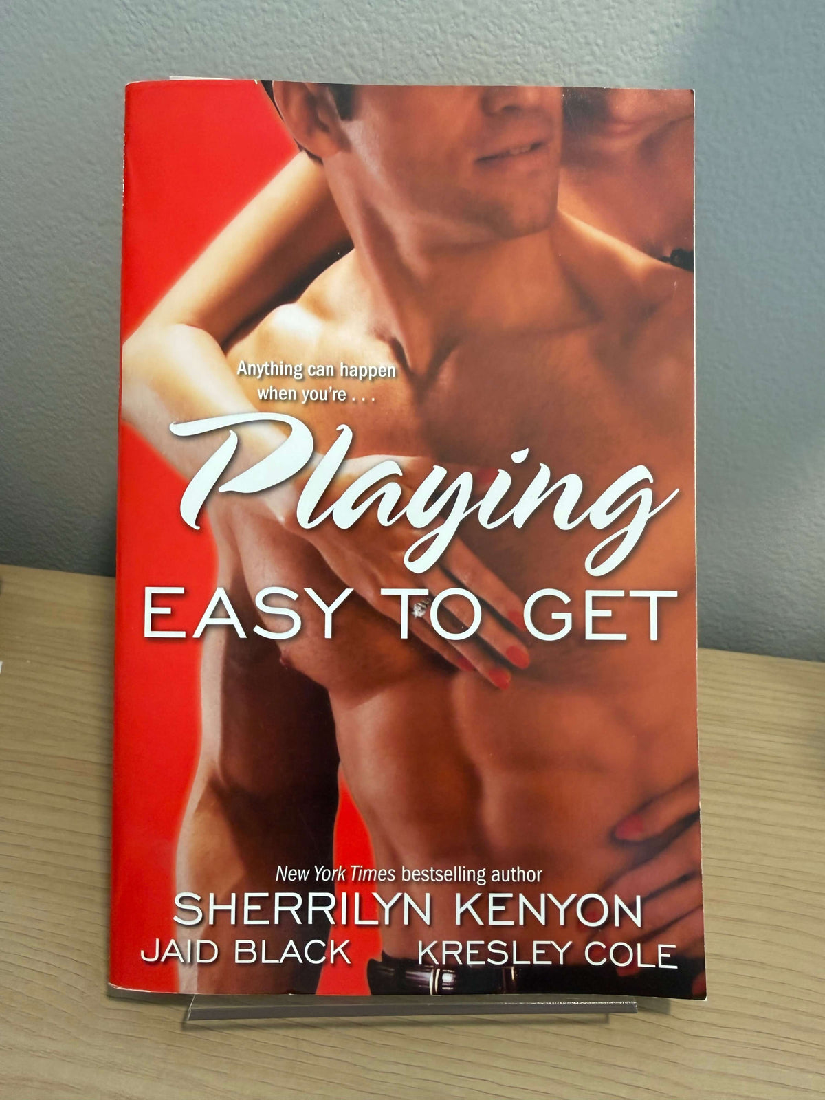 A paperback book titled "Playing Easy to Get" by Sherrilyn Kenyon, Jaid Black, and Kresley Cole, featuring a red cover with a shirtless man on a wooden surface against a gray wall.