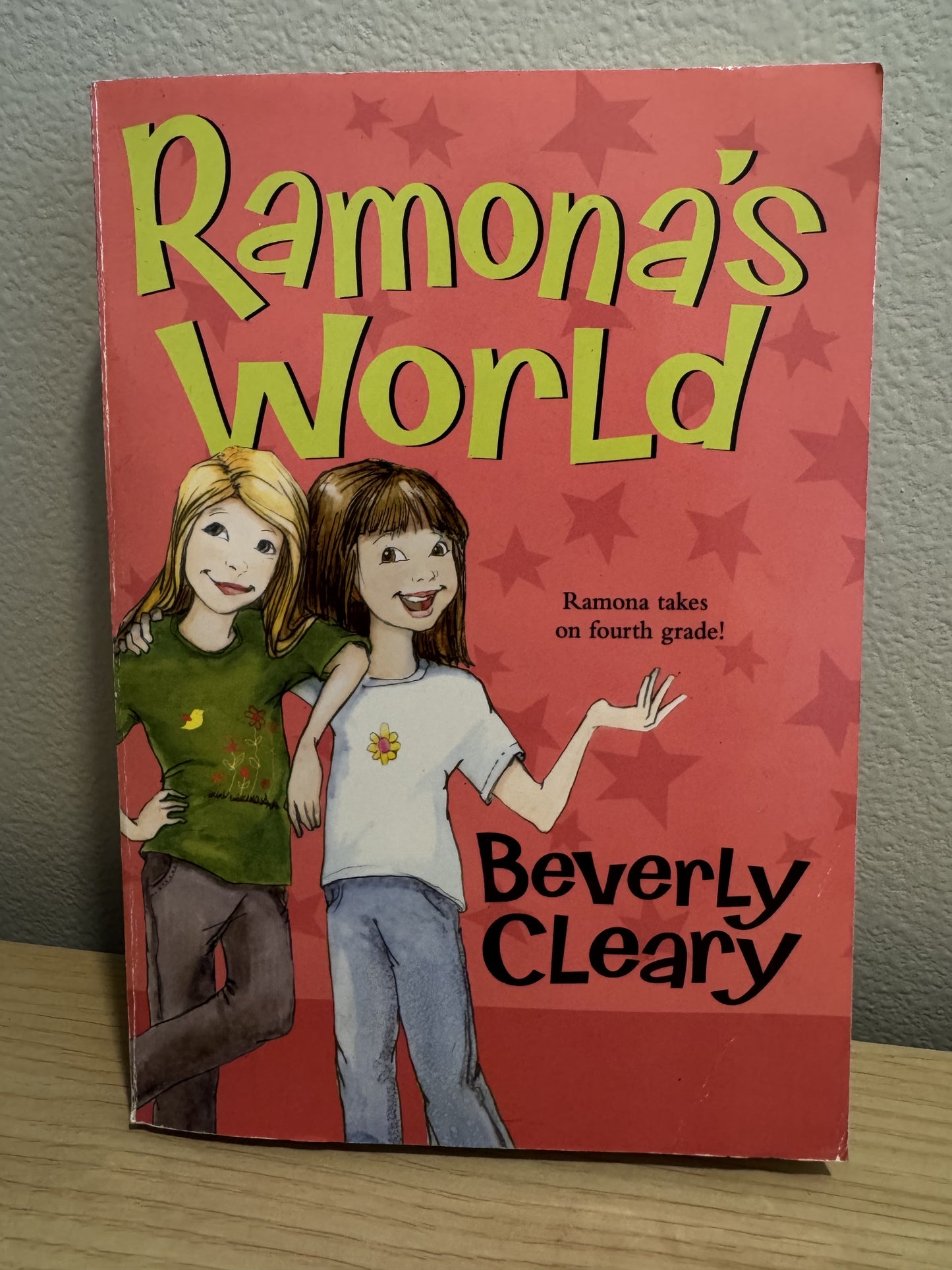 The front cover of the children's book 'Ramona's World' by Beverly Cleary, featuring a red background with star patterns and cartoon illustrations of two girls, shown standing on a wooden surface against a gray wall.