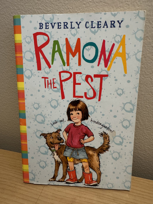 The front cover of the children's book 'Ramona the Pest' by Beverly Cleary, standing upright on a wooden surface against a textured wall. The cover features a light blue-gray background with a pattern of spiderwebs, the title in large multicolored letters, and an illustration of a girl in a red shirt and denim shorts standing next to a brown dog.