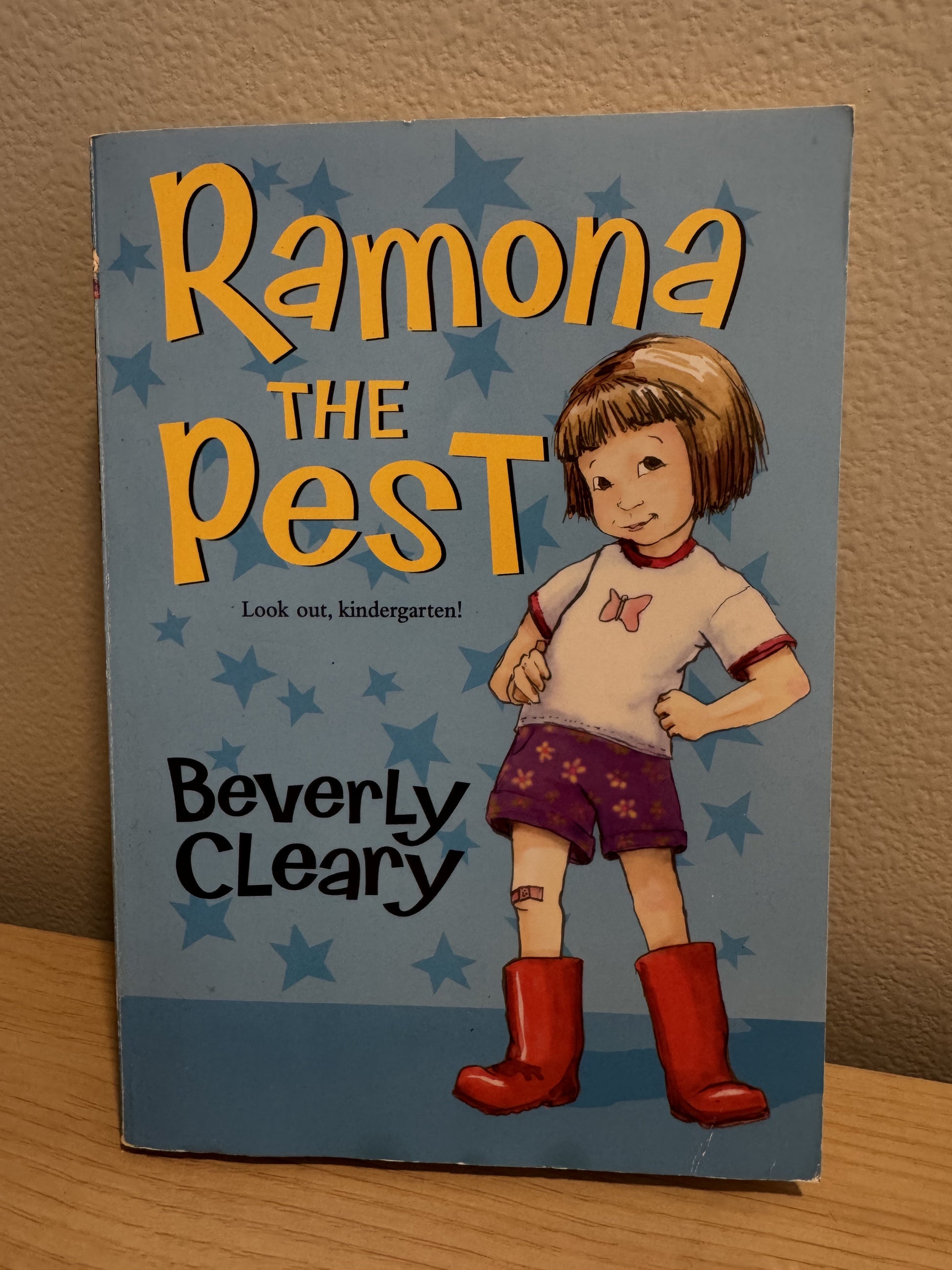 The book cover of 'Ramona the Pest' by Beverly Cleary, featuring a cartoon illustration of Ramona with brown hair, a white t-shirt, purple shorts, and red rain boots on a light blue background with star patterns.