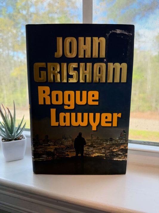 Book titled 'Rogue Lawyer' by John Grisham on a windowsill with a view of trees outside.