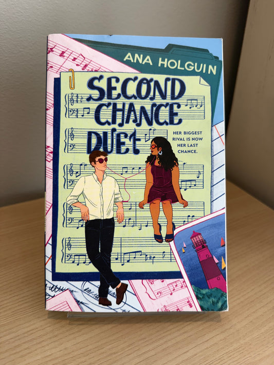 The book cover of 'Second Chance Duet' by Ana Holguin, displayed on a light wood surface with a neutral background. The cover features a colorful illustration of two young adults in front of a sheet music background, with a lighthouse illustration in the bottom right corner.