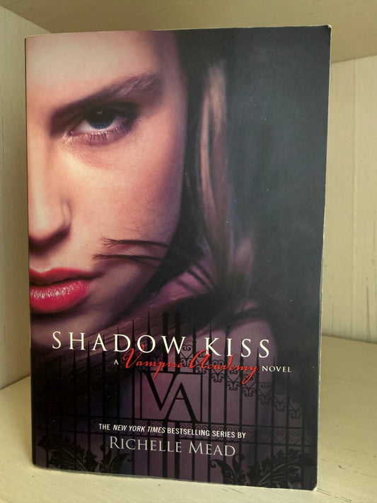 Book cover of 'Shadow Kiss' by Richelle Mead, featuring a close-up portrait of a woman with red lips and a dark gothic background, photographed standing upright on a light-colored surface.