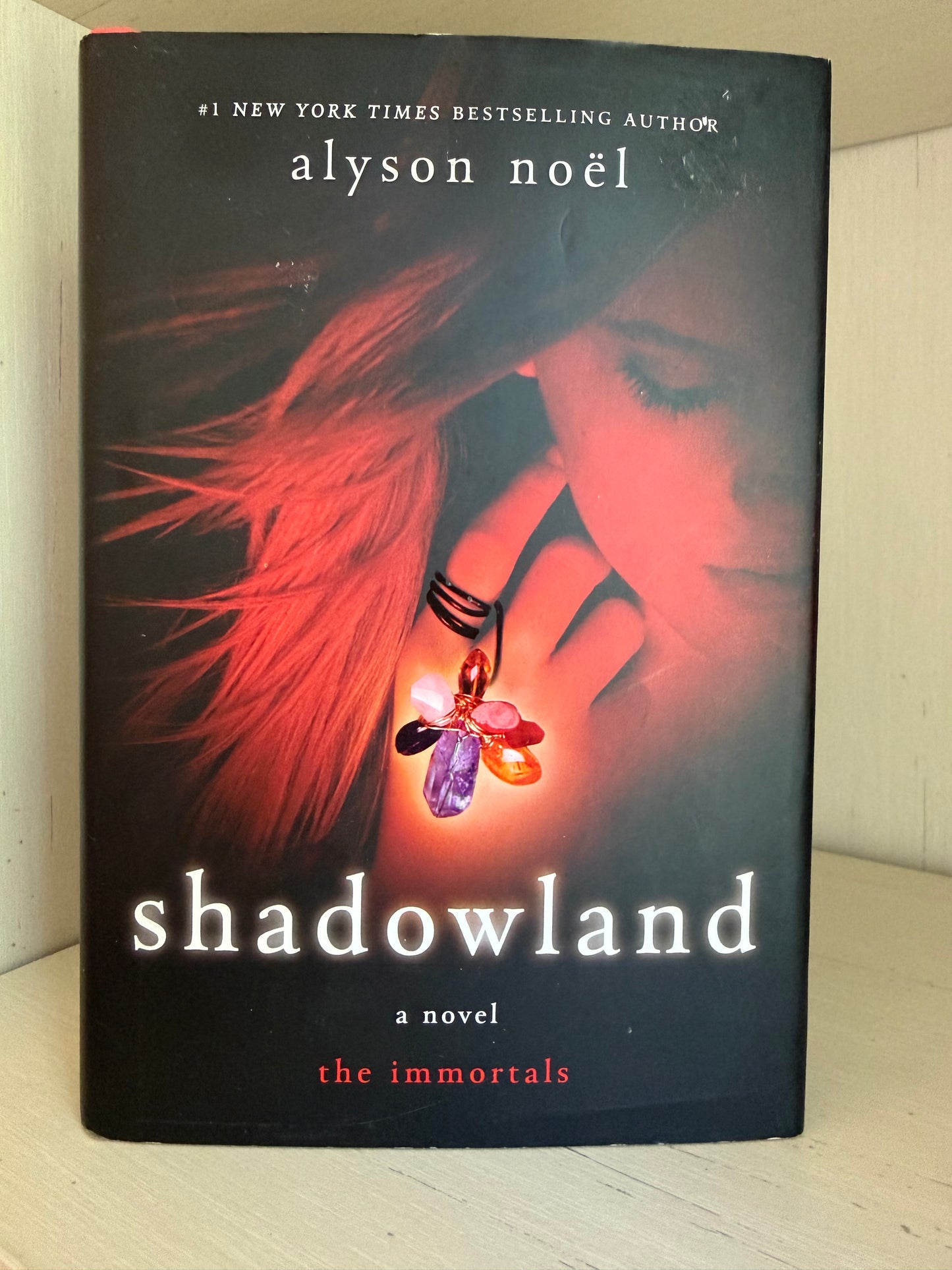 The book cover of 'Shadowland' by Alyson Noel, featuring a dark background with a woman's face in red light, flowing red hair, and a glowing multicolored crystal pendant. The title 'shadowland' is printed in large white letters at the bottom.