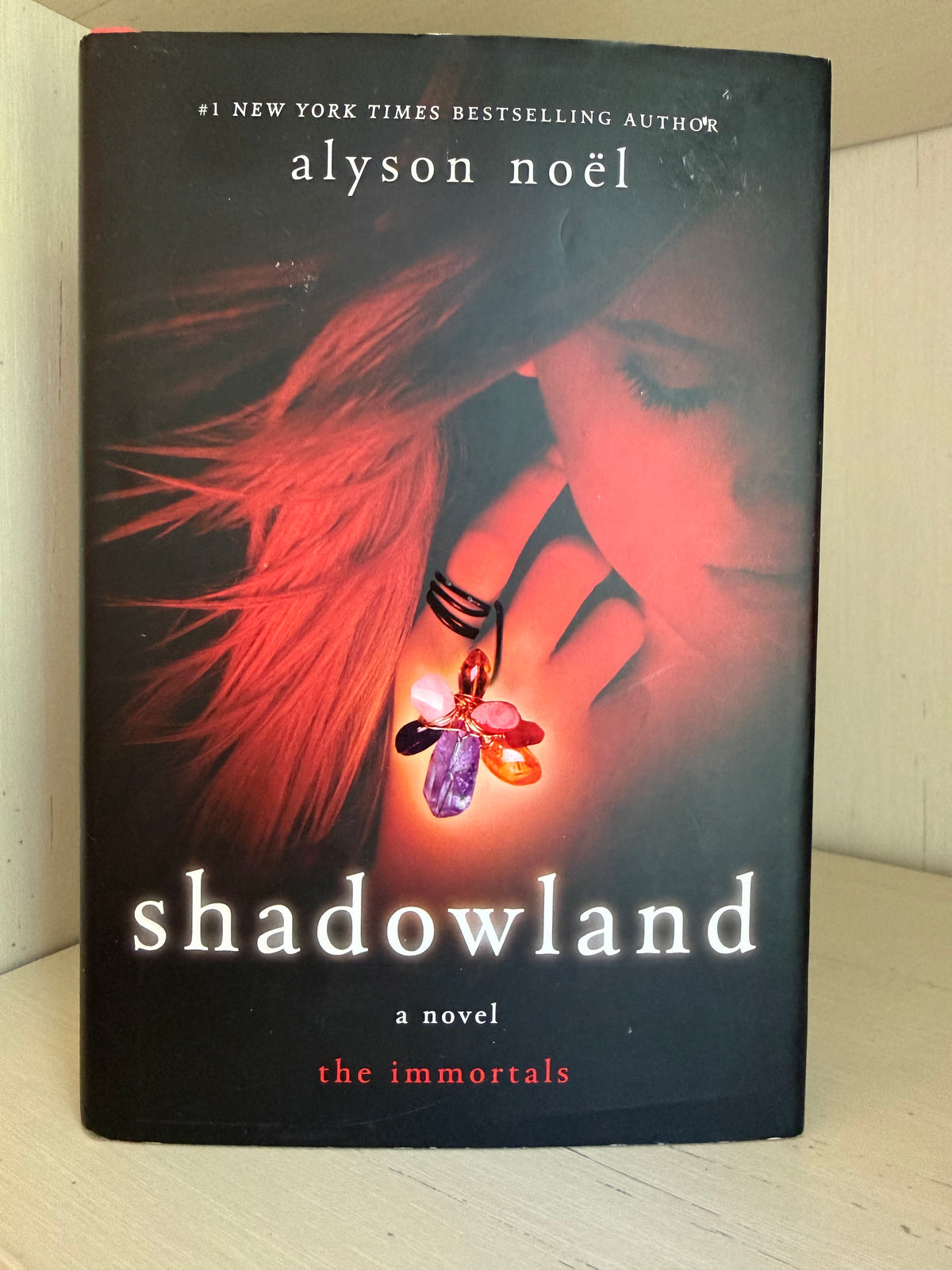 The book cover of 'Shadowland' by Alyson Noel, featuring a dark background with a woman's face in red light, flowing red hair, and a glowing multicolored crystal pendant. The title 'shadowland' is printed in large white letters at the bottom.