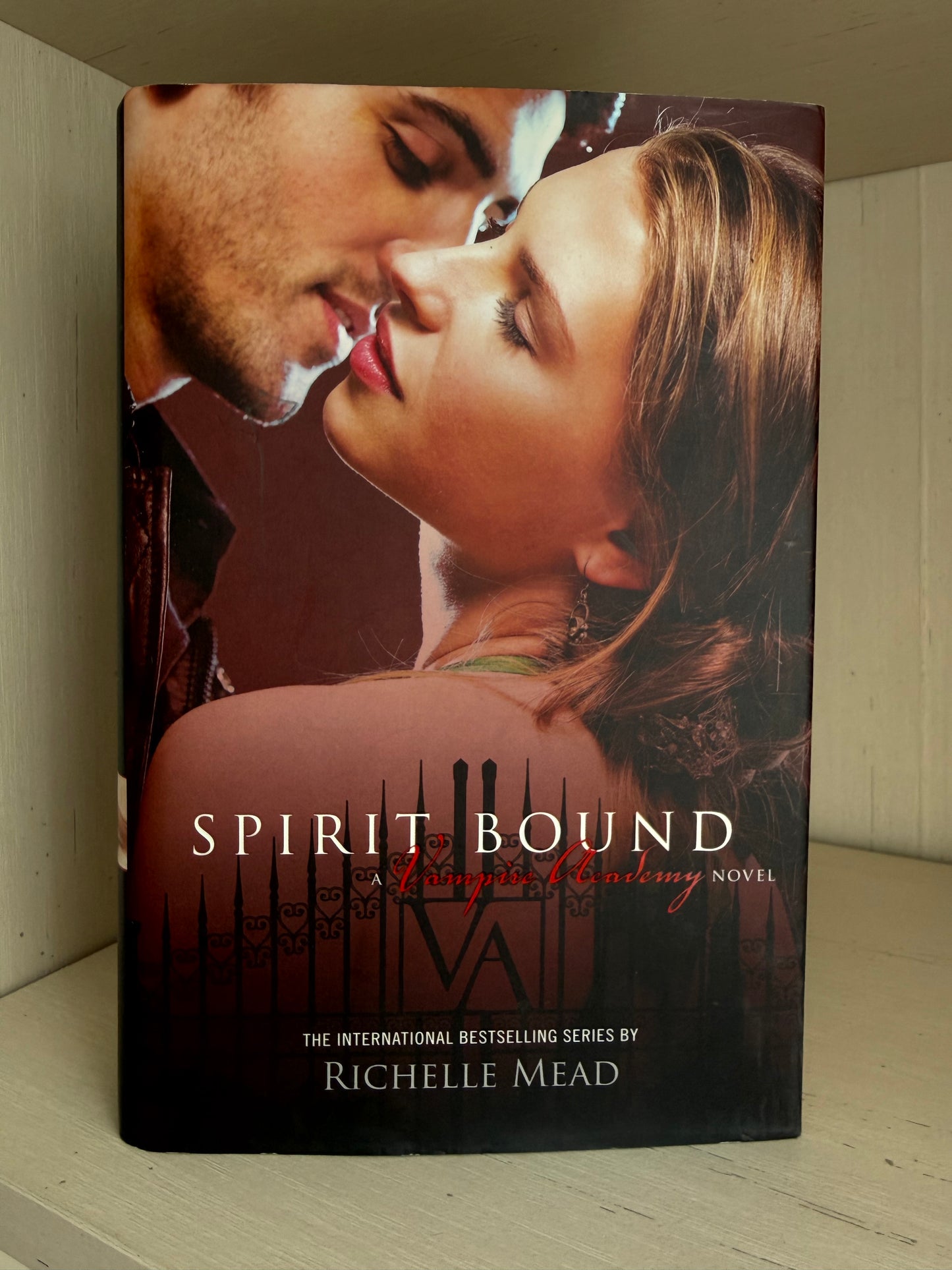 A hardcover book titled 'Spirit Bound: A Simple Romance Novel' by Richelle Mead, standing upright on a light-colored wooden shelf. The cover features a romantic close-up of a man and woman about to kiss, with warm brown and red tones.