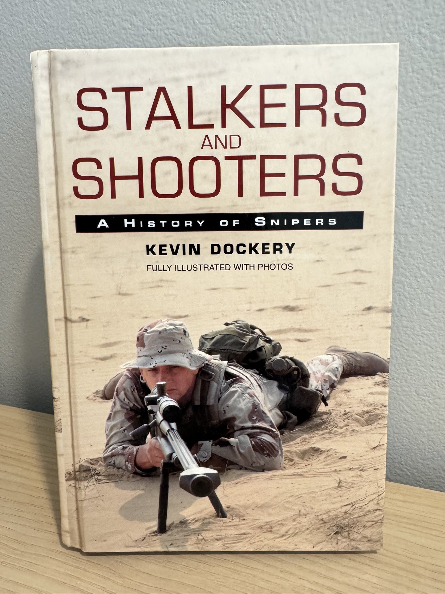The book 'Stalkers and Shooters: A History of Snipers' by Kevin Dockery, standing upright on a light wood surface against a gray wall. The cover features a photograph of a soldier in camouflage lying in sand and aiming a sniper rifle.