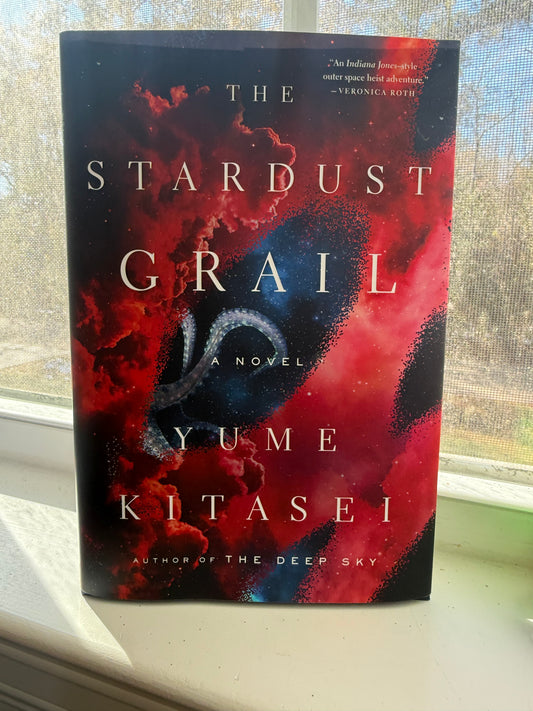 A book titled 'The Stardust Grail' by Yume Kitasei, featuring a vibrant cover with a blend of red and purple hues, placed on a windowsill.