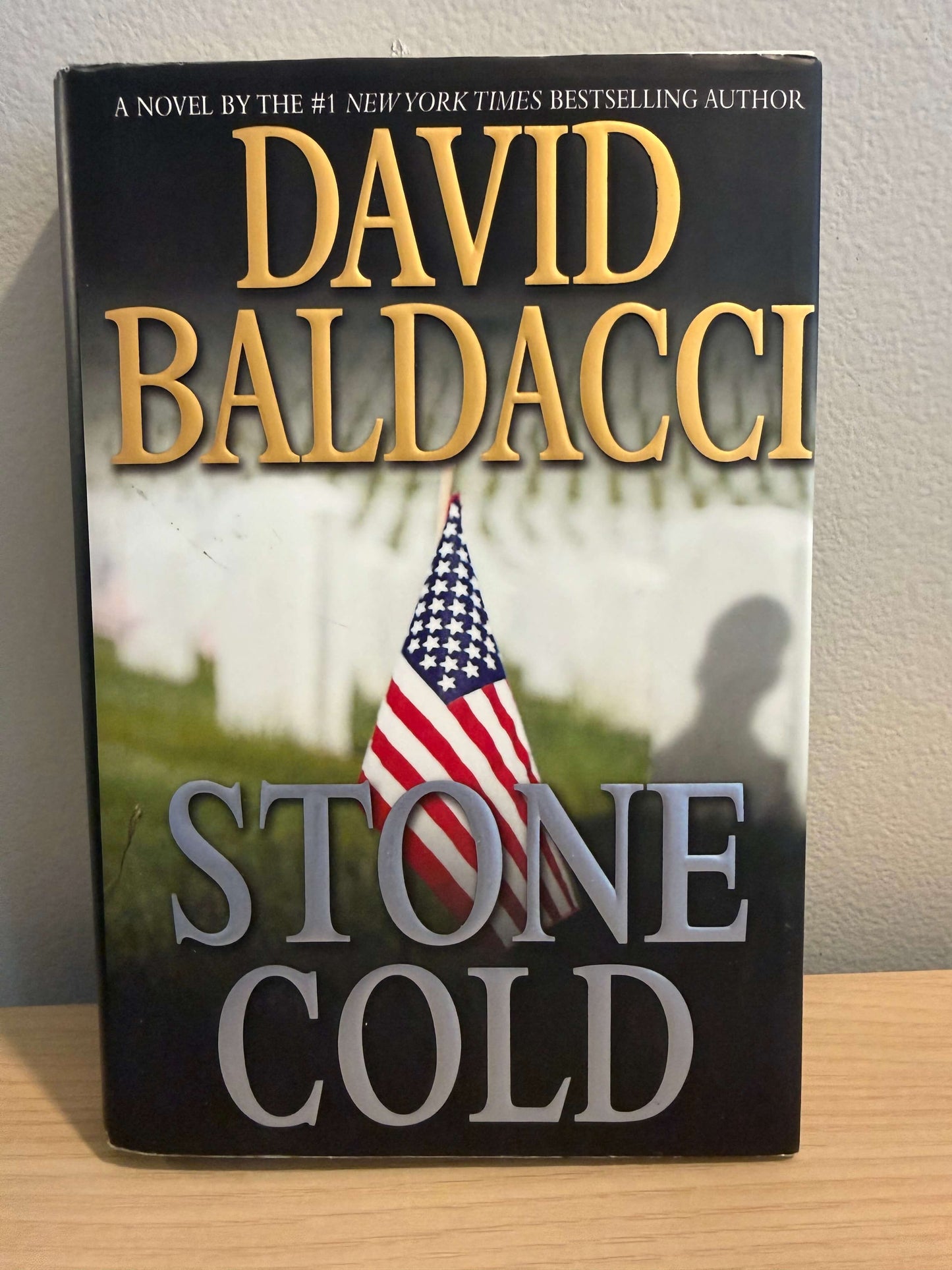 The front cover of the novel 'Stone Cold' by David Baldacci, featuring a black background with gold and gray lettering and an American flag graphic, photographed standing upright on a wooden surface against a light wall.