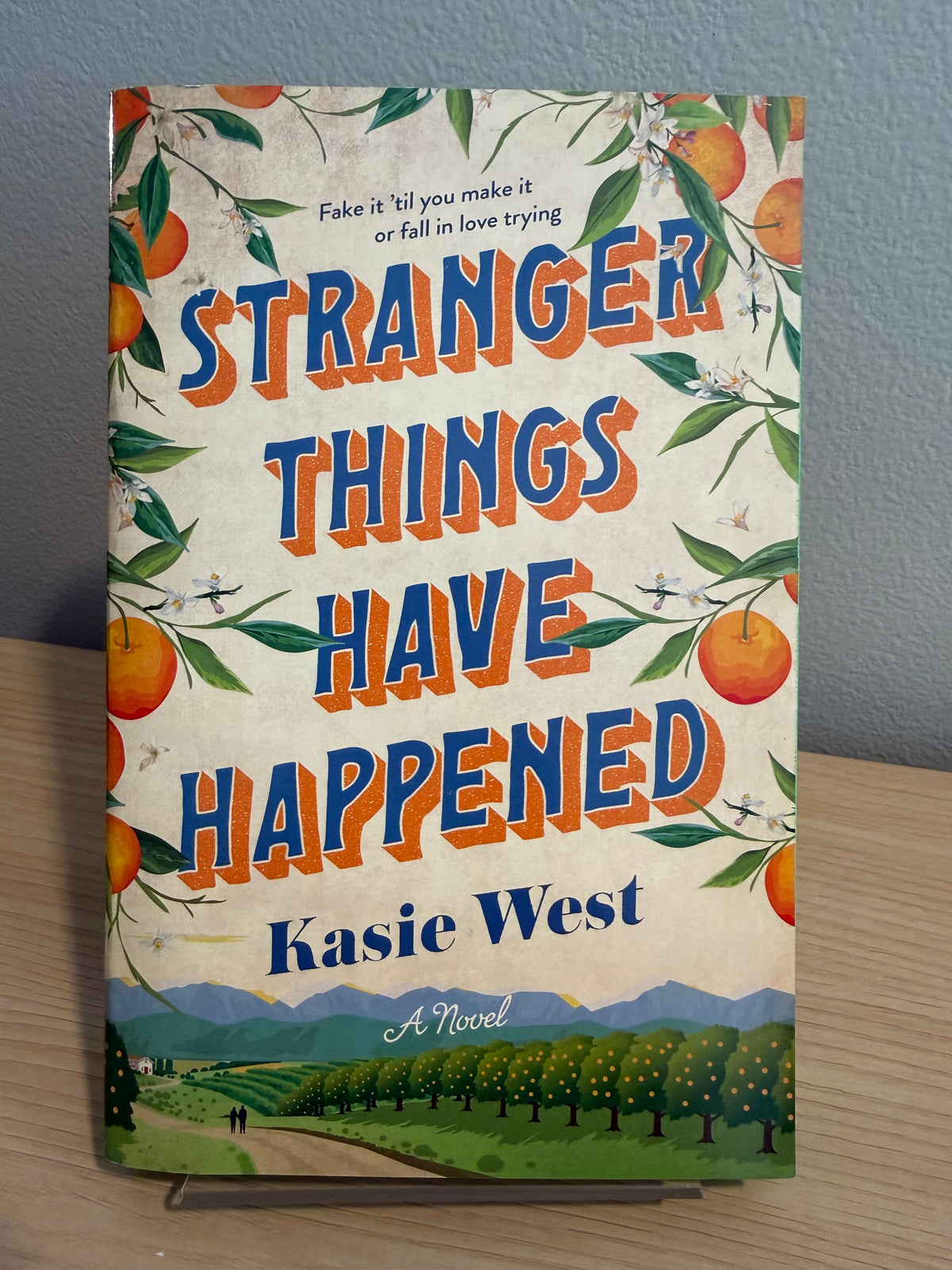 The book cover of 'Stranger Things Have Happened' by Kasie West, featuring a vintage-style illustration of an orange grove and a small landscape scene at the bottom, displayed on a wooden surface against a gray wall.
