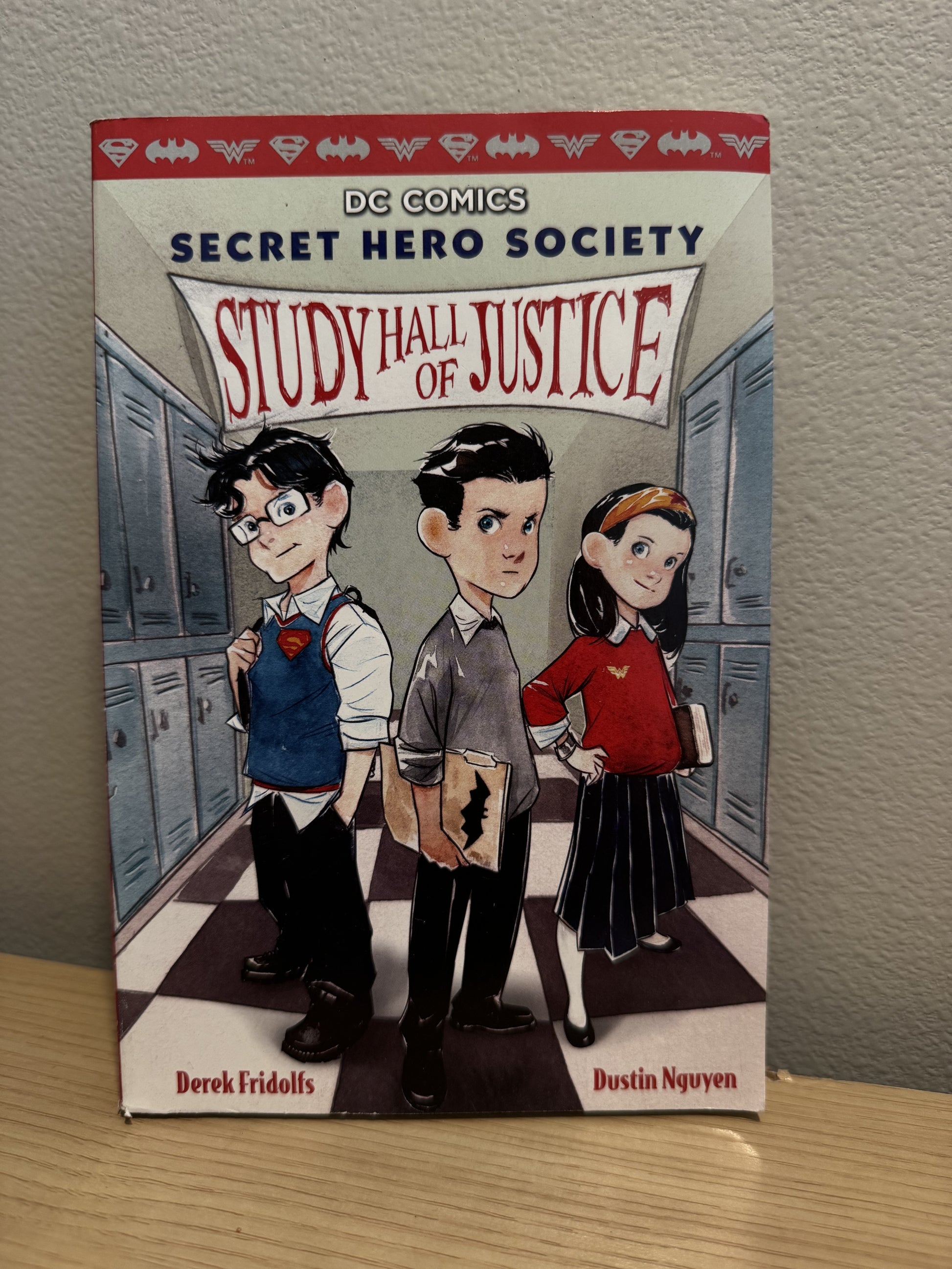 The front cover of the book 'Study Hall of Justice' from DC Comics' Secret Hero Society series, featuring three illustrated young superheroes standing in a school hallway. The book is photographed standing upright on a light wood surface against a textured beige wall.