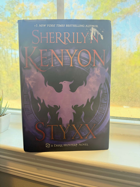 Book titled 'Styxx' by Sherrilyn Kenyon on a windowsill with a blurred outdoor background
