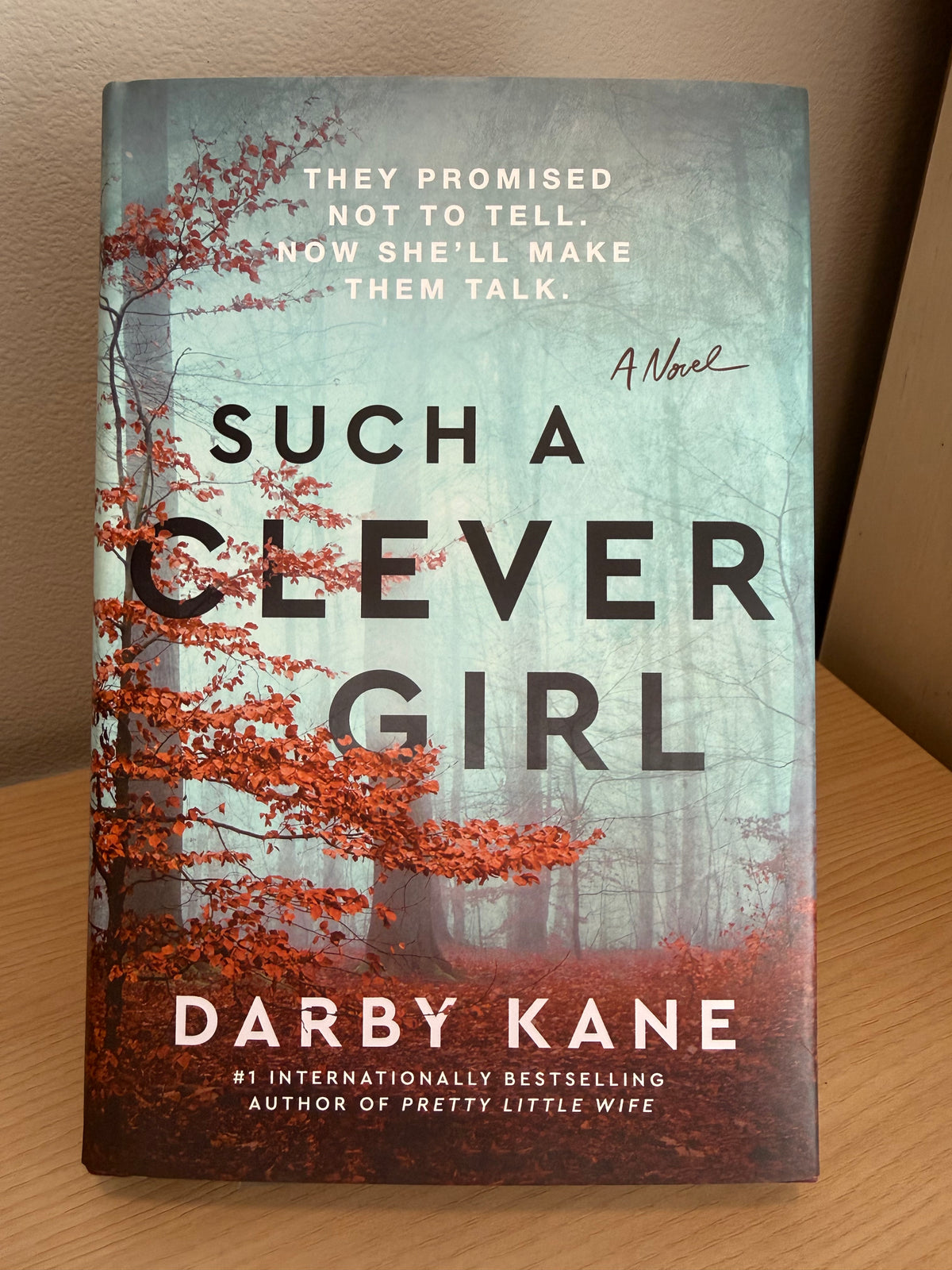 The front cover of the novel 'Such a Clever Girl' by Darby Kane, featuring a misty forest with bright orange-red autumn leaves, photographed standing on a wooden surface against a beige wall.