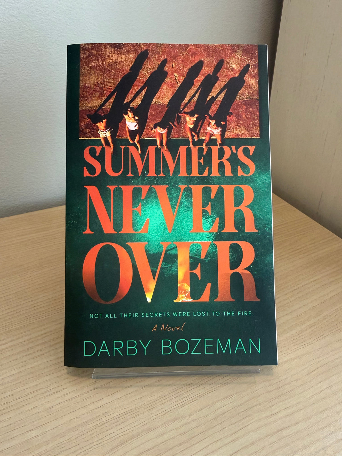 The book 'Summer's Never Over' by Darby Bozeman, standing upright on a clear display stand on a light wood surface against a neutral background.