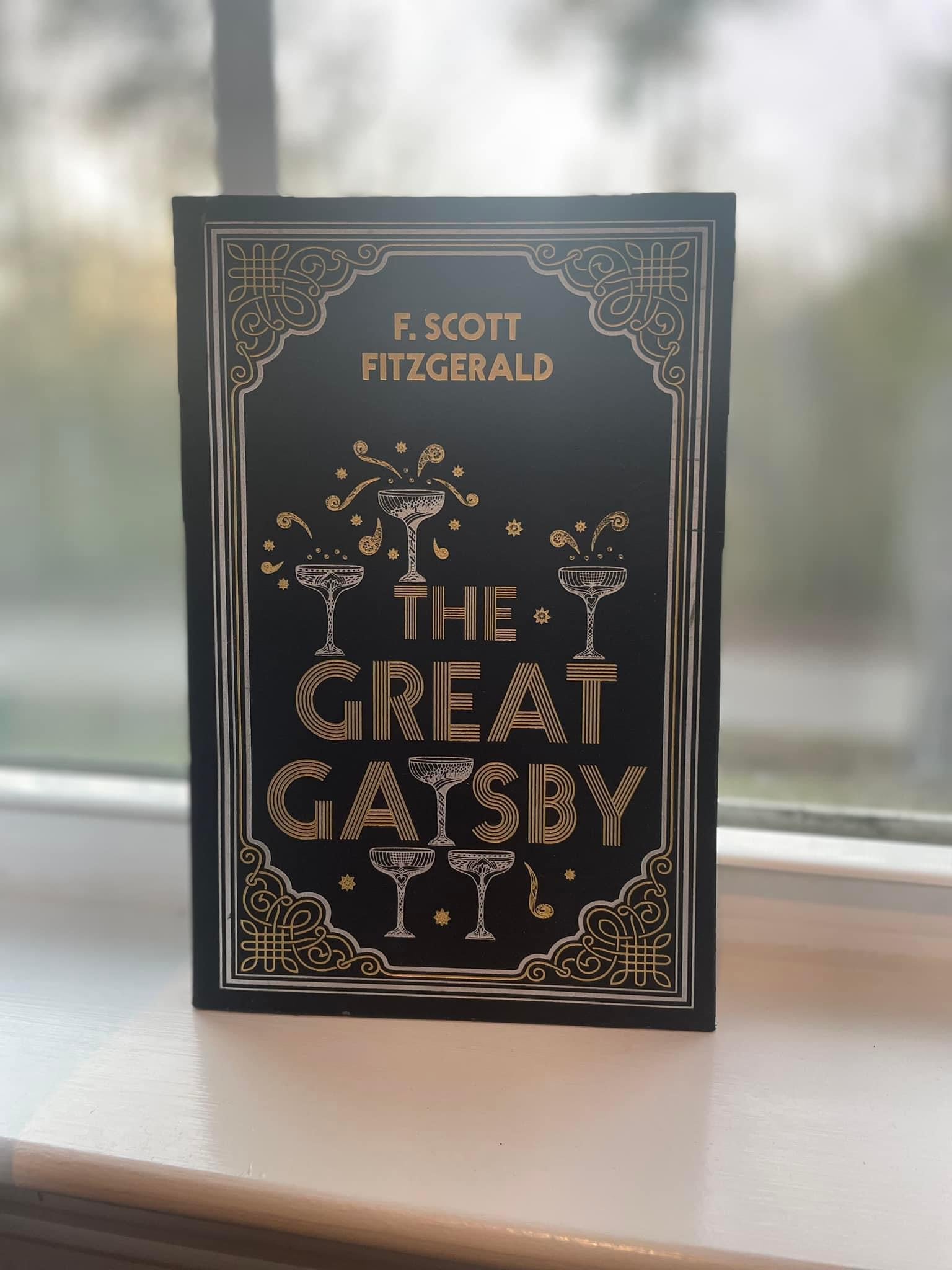 A book titled 'The Great Gatsby' by F. Scott Fitzgerald, with a black and gold cover design, placed on a window sill.