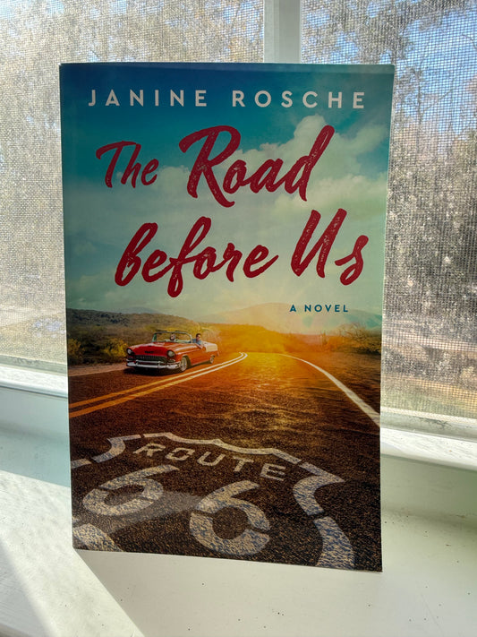 A book cover of 'The Road Before Us' by Janine Rosche, featuring a red and white design with text, Route 66, and a silhouette of a car on the cover.