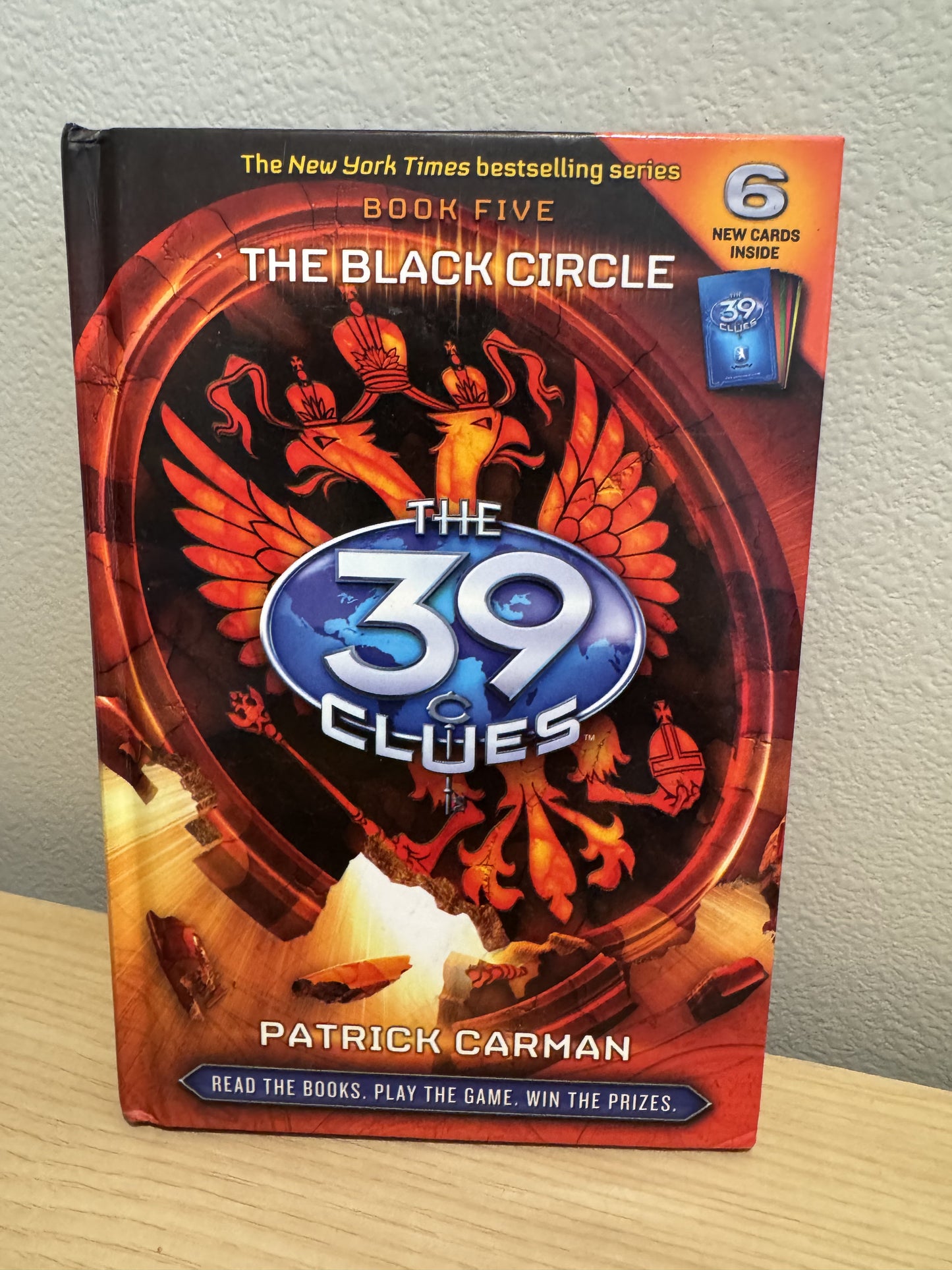 The Black Circle: Book Five from The 39 Clues series by Patrick Carman, featuring a red and orange cover with a blue central logo, standing on a wooden surface against a light gray wall.