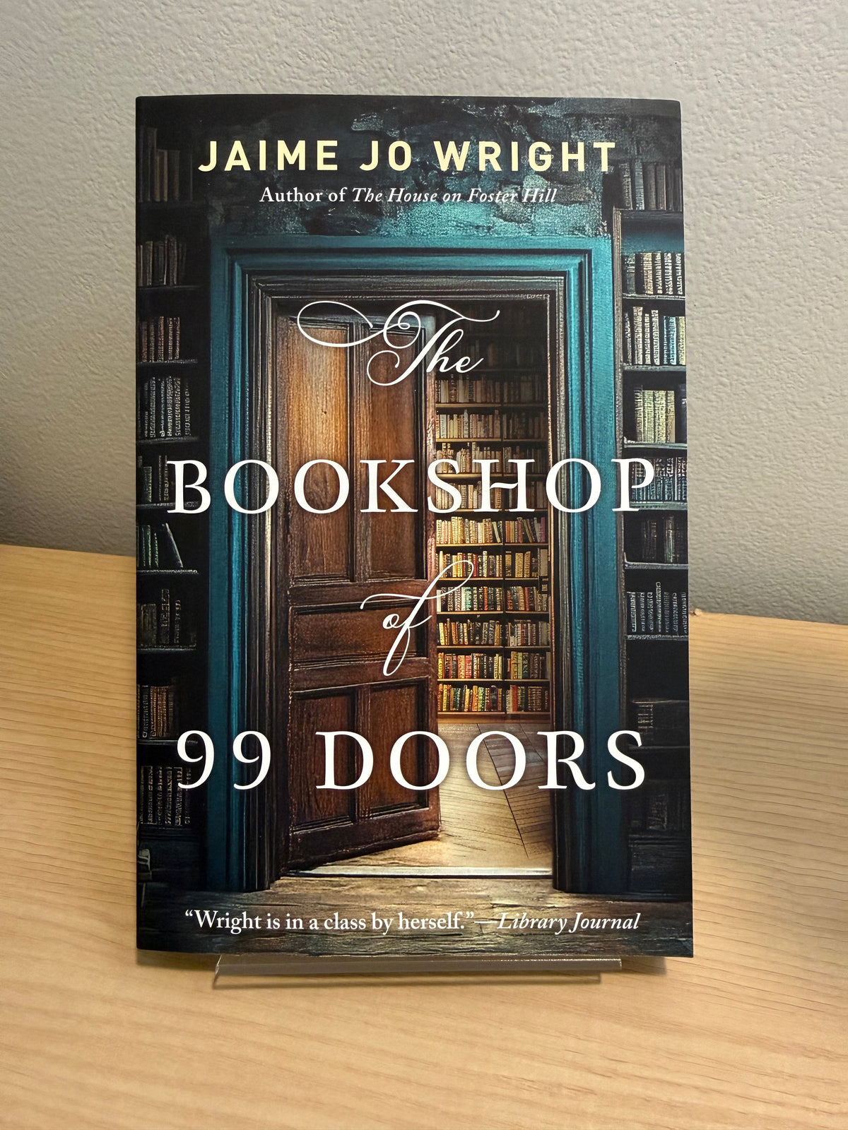 The Bookshop of 99 Doors paperback book by Jaime Jo Wright, standing upright on a small clear display stand on a light wood surface against a neutral wall.