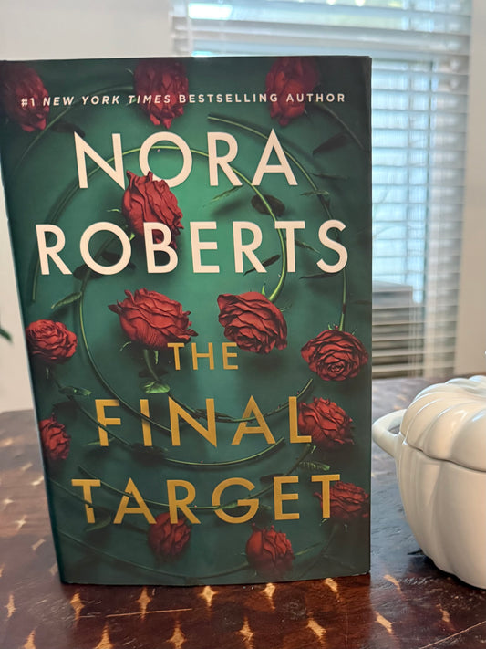 The book cover of Nora Roberts' 'The Final Target' is shown standing upright on a wooden table, with a white ceramic container and window blinds in the background.
