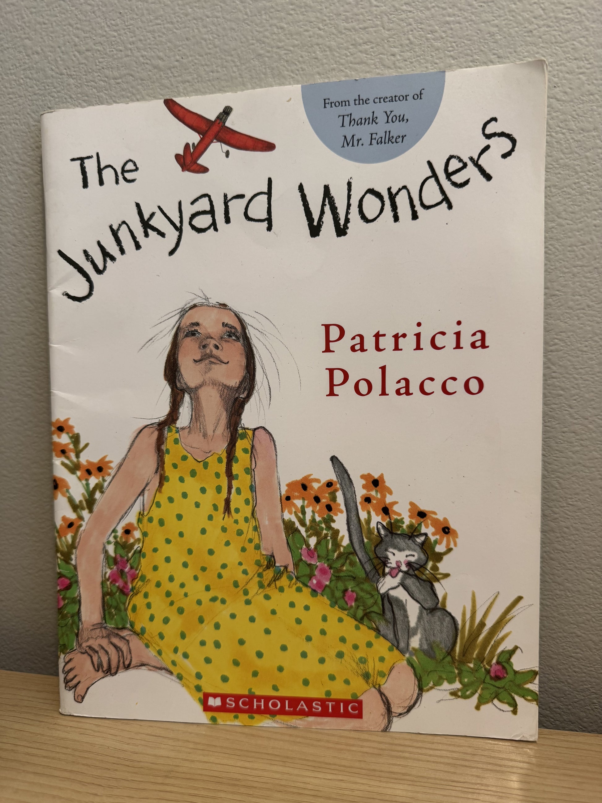 The Junkyard Wonders book by Patricia Polacco, published by Scholastic, featuring a colorful illustration of a girl in a yellow polka-dot dress, a red airplane, and a cat on the cover, photographed standing on a wooden surface against a gray wall.