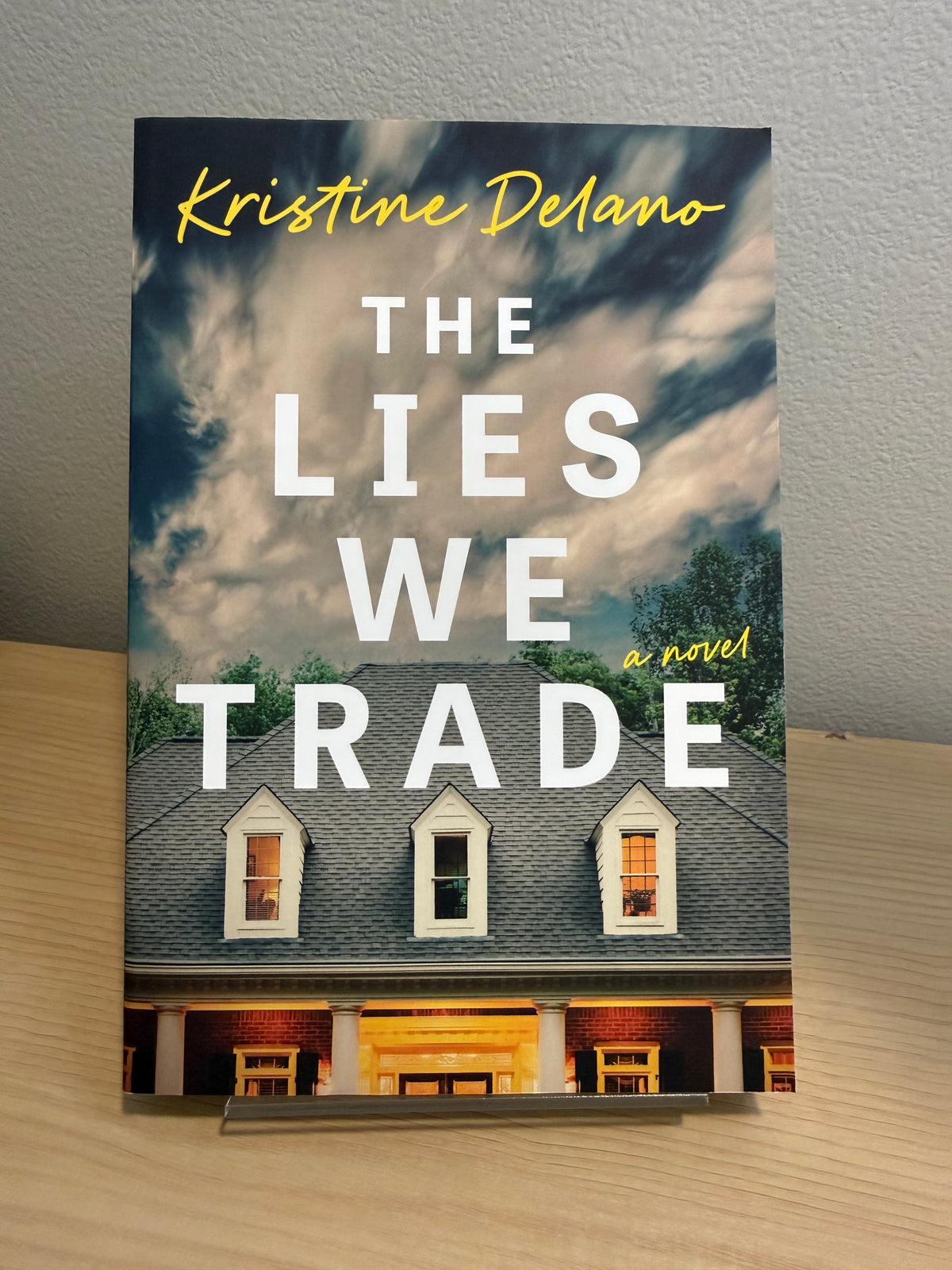 The front cover of the novel 'The Lies We Trade' by Kristine Delano is displayed standing upright on a small clear acrylic display block on a light wood surface against a textured gray wall.