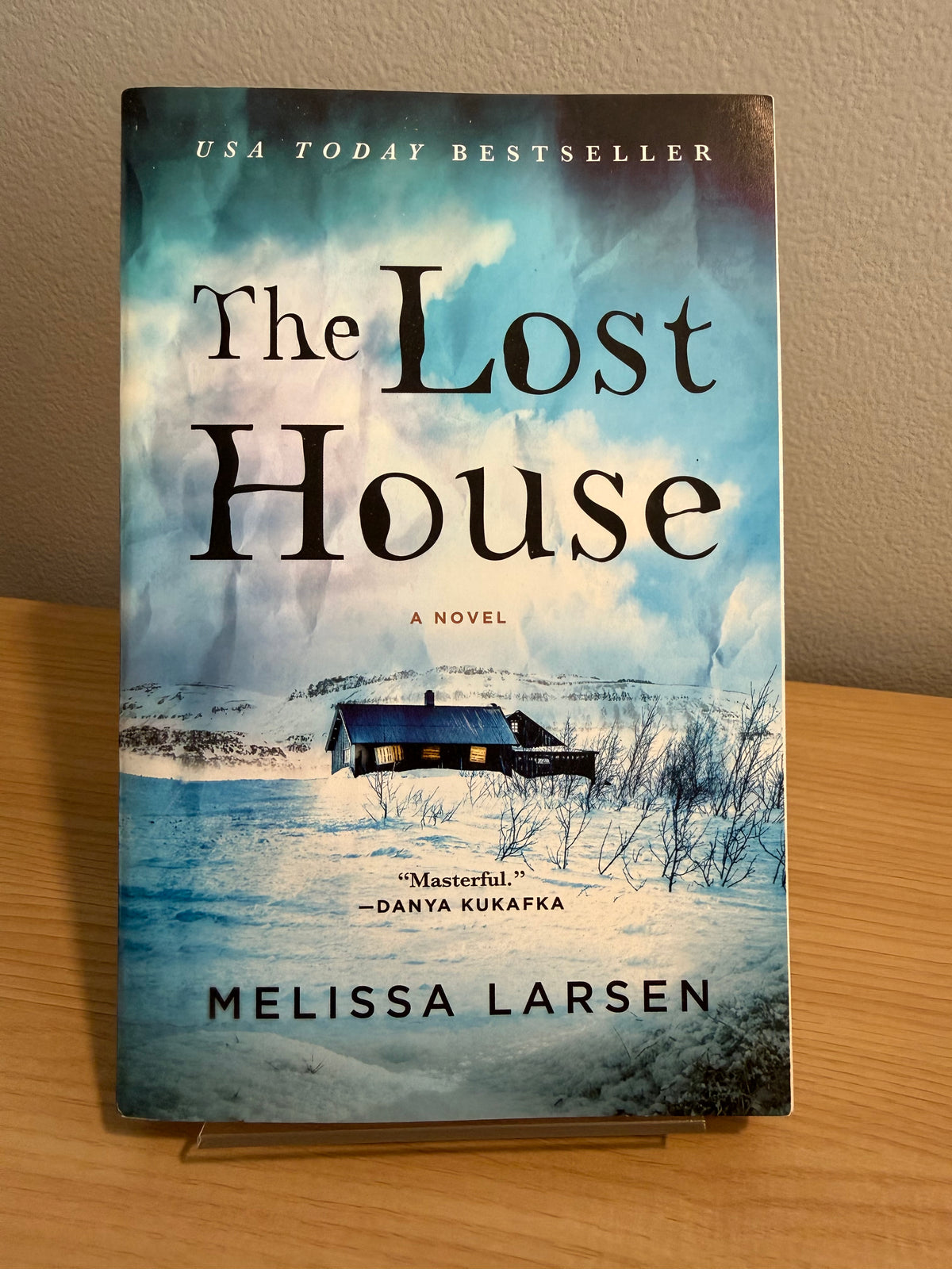 The Lost House: A Novel by Melissa Larsen, standing upright on a small clear display stand on a wooden table against a beige wall.