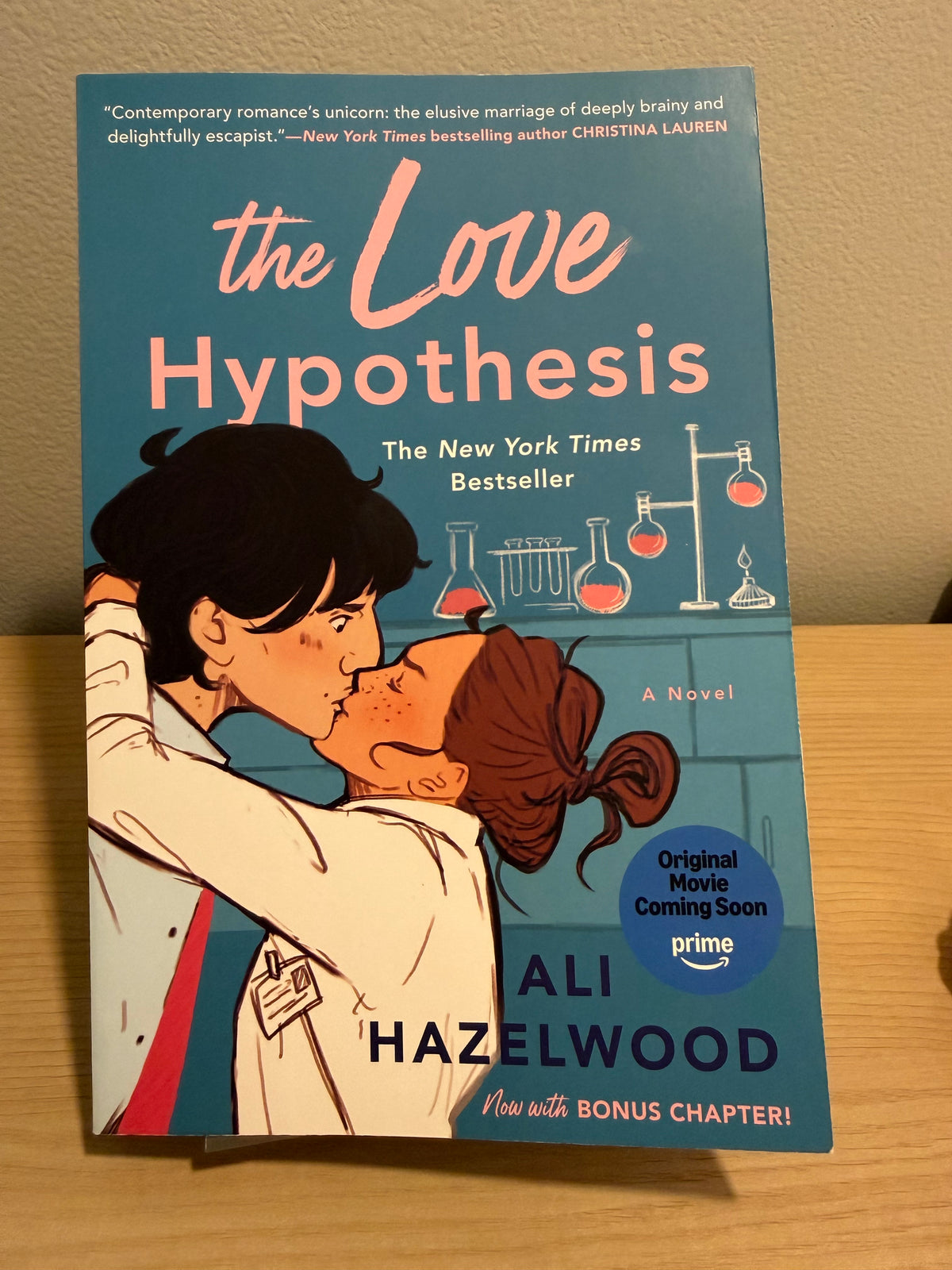 The Love Hypothesis book cover by Ali Hazelwood, featuring an illustration of a couple in lab coats with chemistry equipment on a teal background, photographed standing on a wooden surface against a beige wall.
