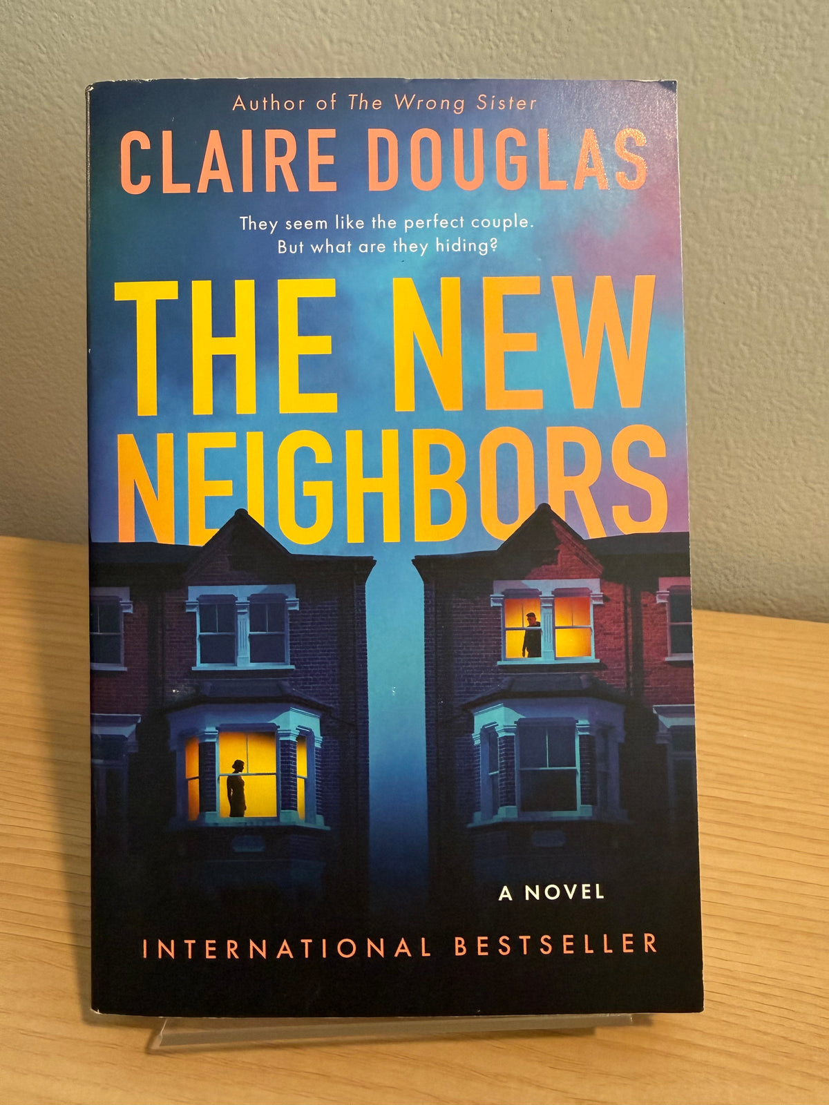 The New Neighbors paperback novel by Claire Douglas, featuring a dark blue and purple night sky background with two houses and glowing yellow windows, photographed standing upright on a small clear display against a beige wall and wood surface.