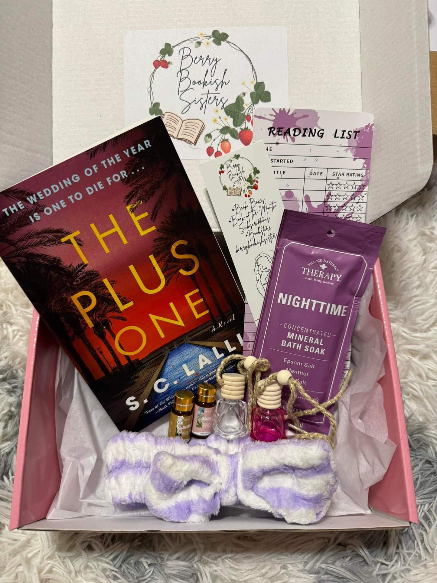 Gift box with 'The Plus One' book, bath salts, and other items on a textured surface.