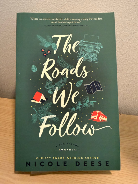 The front cover of the book 'The Roads We Follow' by Nicole Deese, featuring a dark teal-green background with cream lettering and small illustrated accents, photographed standing on a light wood surface against a beige wall.