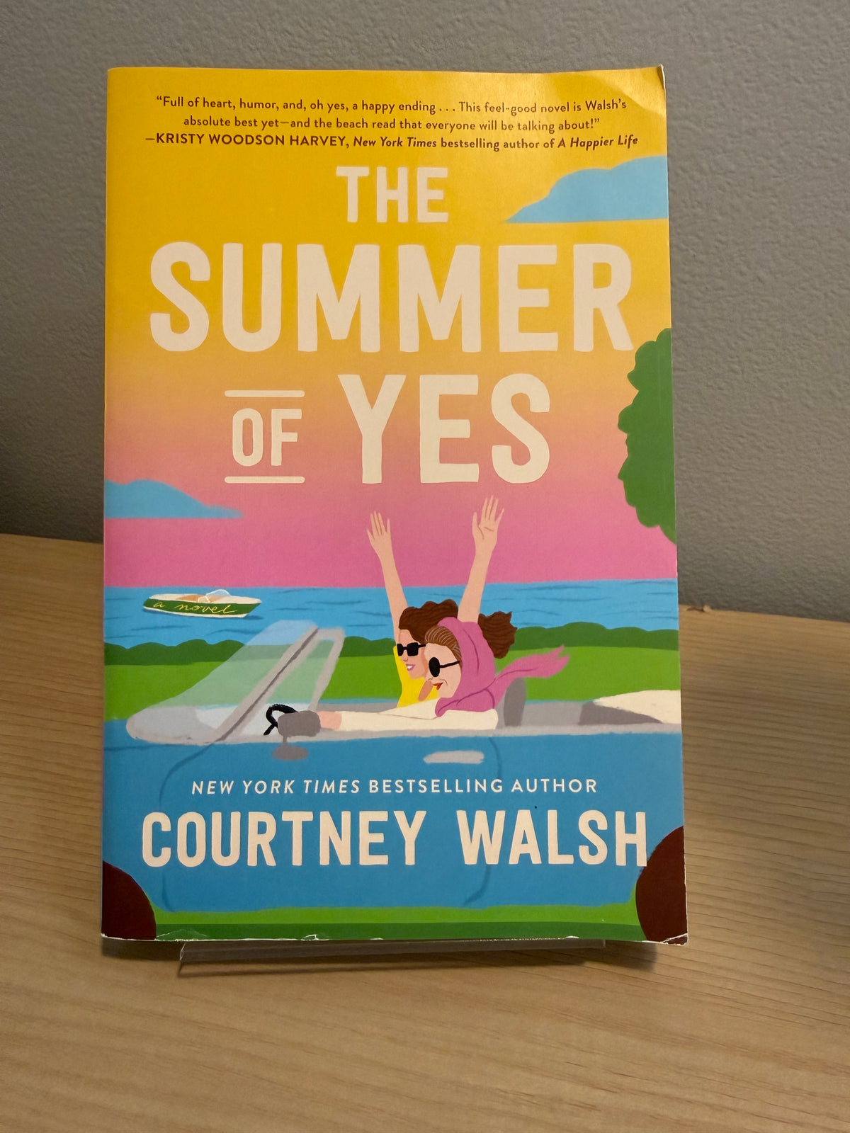 The Summer of Yes paperback book by Courtney Walsh, standing upright on a small display stand on a wooden surface against a gray wall.