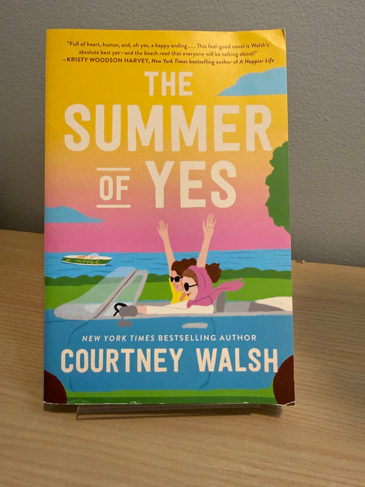 The Summer of Yes paperback book by Courtney Walsh, standing upright on a small display stand on a wooden surface against a gray wall.