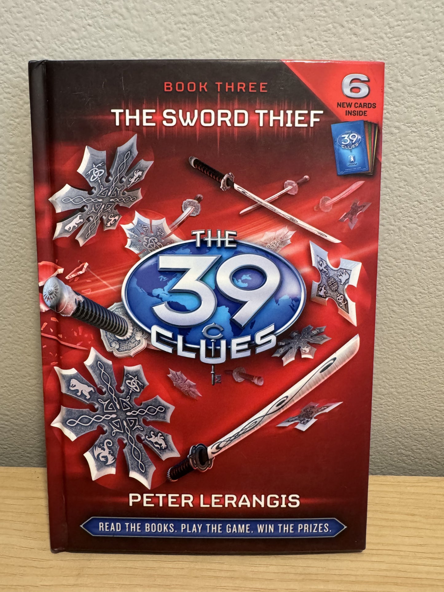 The Sword Thief book 3 from The 39 Clues series by Peter Lerangis, featuring a red cover with silver and blue elements, standing on a wooden surface against a beige wall.