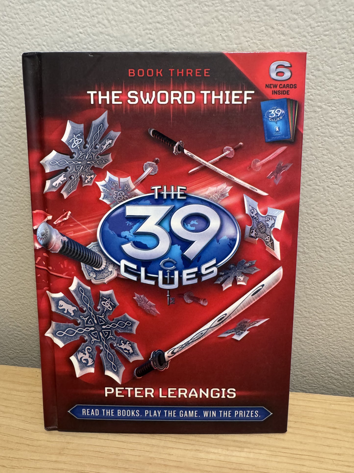 The Sword Thief: The 39 Clues Book 3 board game by Peter Lerangis, featuring a red and black cover with silver swords and snowflake designs, displayed on a wooden surface against a beige wall.