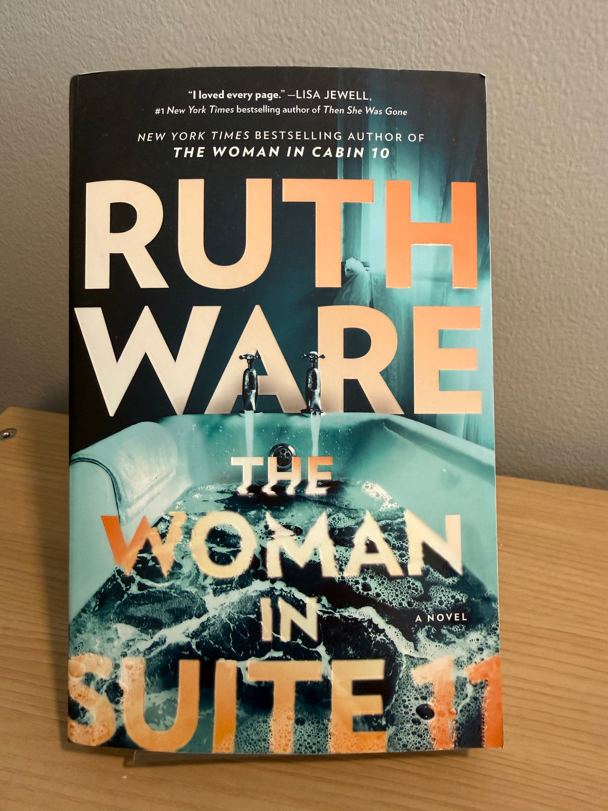 The book cover of 'The Woman in Suite 11' by Ruth Ware, photographed standing upright on a wooden surface against a beige wall.