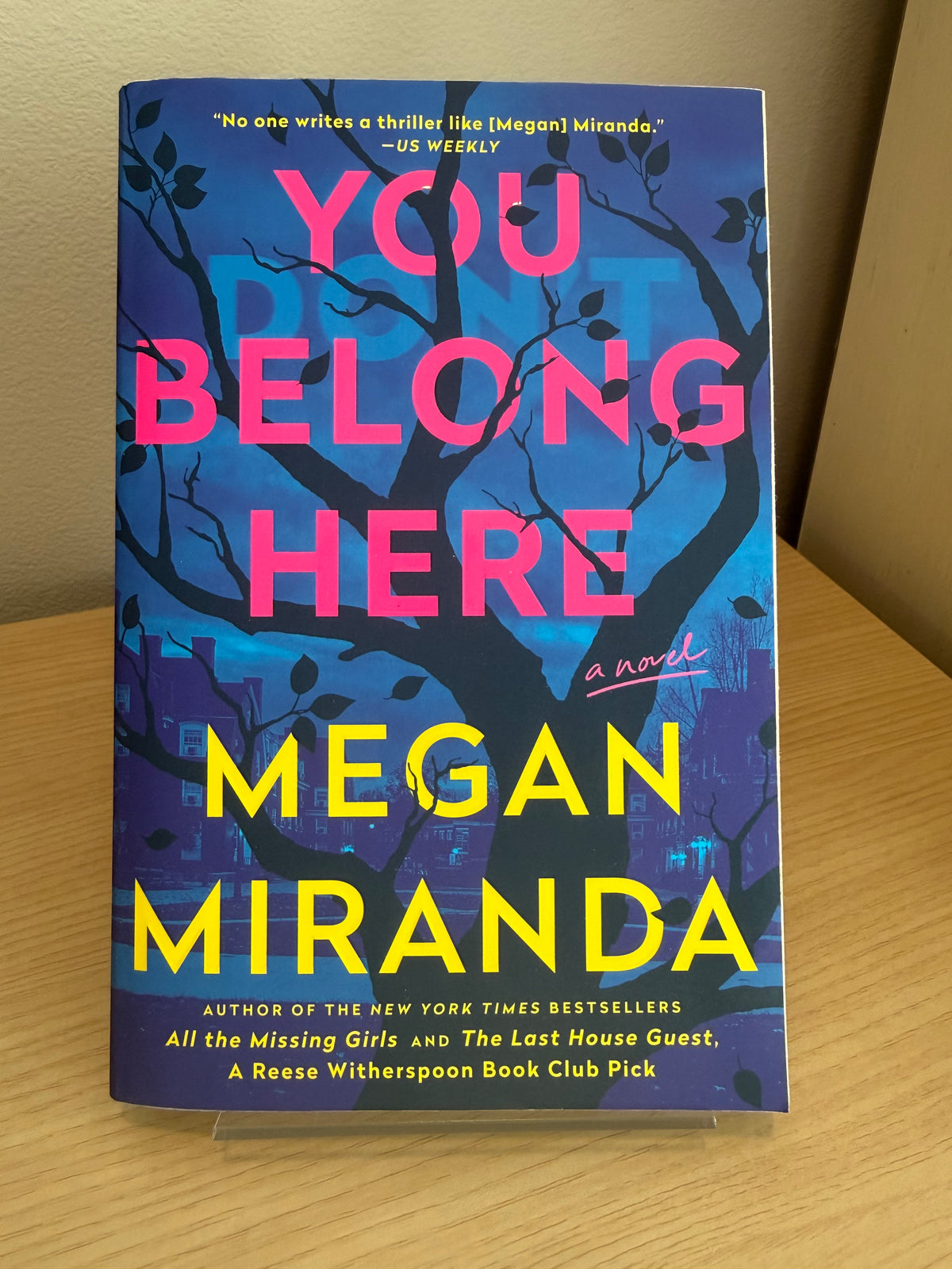 The book 'You Belong Here' by Megan Miranda, featuring a dark blue and purple cover with pink and yellow text, displayed on a wooden surface.